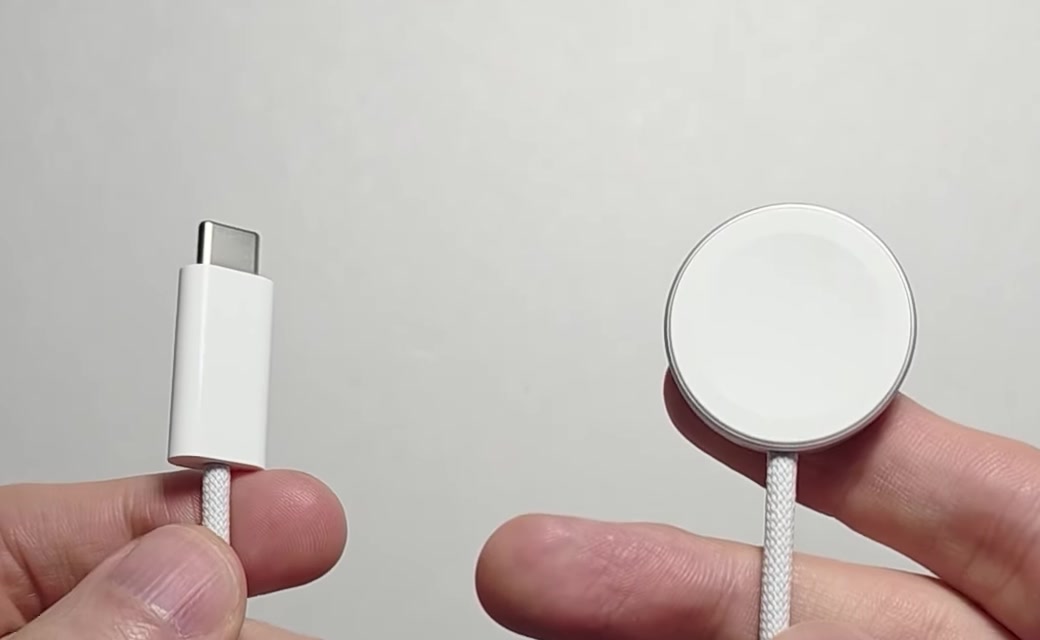 Apple Watch magnetic charging cable held up, showing the USB-C connector at one end and the round magnetic puck at the other