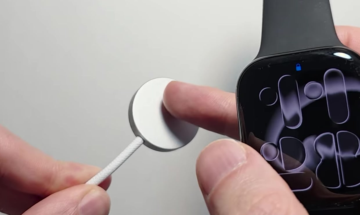 The magnetic charging puck being brought up to the back of an Apple Watch, which is lying face-up on a desk